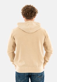 Felpa in cotone beige con cappuccio regolabile, polsini e orlo a coste. Il tessuto ha una consistenza liscia con una finitura leggermente consumata.