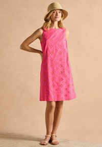 Rosa ärmelloses Kleid mit floralen Stickereien, knielang, aus leichtem Stoff. Modell trägt beige Sandalen und einen Strohhut.