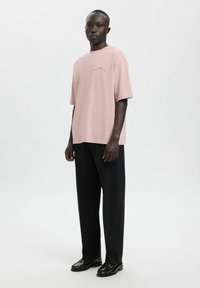 Jasnoróżowy bawełniany t-shirt z krótkimi rękawami i okrągłym dekoltem, noszony z czarnymi szerokimi spodniami i czarnymi butami, stojący na szarym tle.