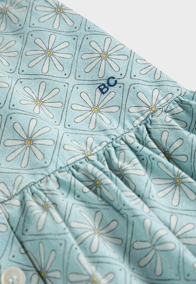 Svetlomodrá tkanina s motívom margarét v bielej a žltej farbe, obsahujúca malé štvorcové obrysy a záhyby blízko vyšívaných písmen "BC".