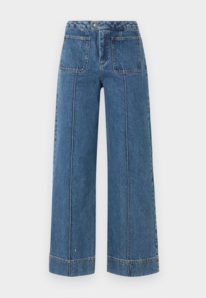 Vrouw draagt een oversized wit overhemd, hooggesneden wijde blauwe spijkerbroek met voorzakken, zwarte ballerina’s en een bruine zonnebril.