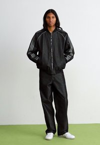 Chaqueta bomber negra con cremallera plateada y detalles a rayas, combinada con pantalones negros. El modelo lleva zapatillas blancas sobre un fondo verde.