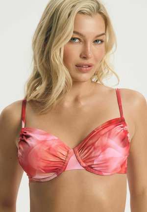 Blond žena s modrými očami nosi ružovo-červený kvetinový bikini top, pózuje na svetlom pozadí.