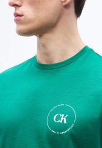 Νεαρός άνδρας που φοράει πράσινο t-shirt Calvin Klein με λευκό λογότυπο CK και κυκλικό κείμενο στην αριστερή πλευρά του στήθους, εμφανιζόμενος από τον λαιμό έως το μέσο του κορμού.