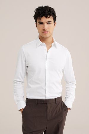 Bărbat cu păr creț și închis la culoare, purtând o cămașă albă cu nasturi și pantaloni maro, stând cu mâinile în buzunare pe un fundal simplu.