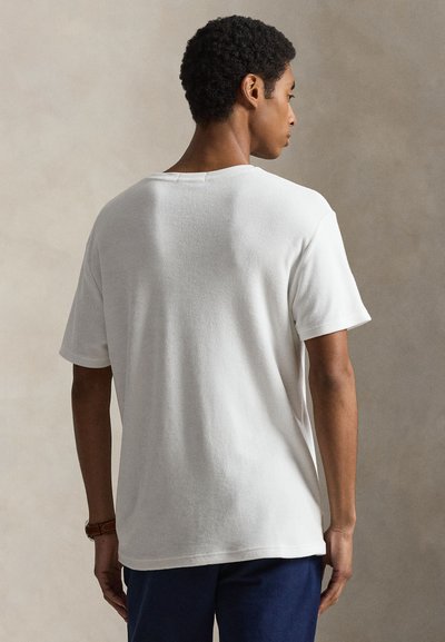 Άνδρας με κοντά σγουρά μαλλιά, φορώντας ένα απλό λευκό κοντομάνικο μπλουζάκι και σκουρόχρωμο παντελόνι, με θέα από πίσω σε ουδέτερο φόντο.