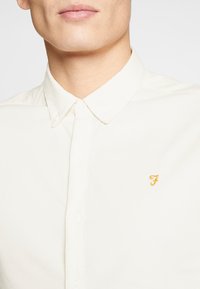Kremowa koszula z kołnierzykiem, wykonana z lekkiego materiału, z małym żółtym wyszywanym logo po lewej stronie klatki piersiowej, z zapięciem na guziki z przodu i gładką teksturą.