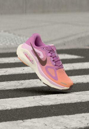 Lila és narancssárga Nike futócipő levegőben, egy fekete-fehér csíkos zebra fölött, talpa alatt por részecskék.