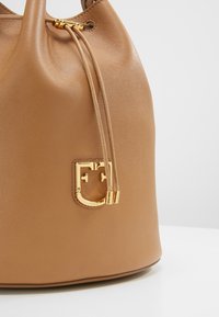 Sac seau en cuir texturé couleur fauve avec cordon de serrage doré et emblème du logo à l'avant sur fond blanc.