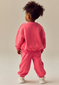 Erksavärvilisest roosast dressipluusi ja pükste komplekt, millel on soonikkoes mansetid ja vöökoht. Pehme kangas ja mugav lõige. Valged tossud täiendavad komplekti.