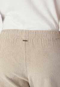 Pantalones de pana beige con una textura acanalada, que cuentan con una cinturilla elástica y una pequeña etiqueta metálica con el logo.