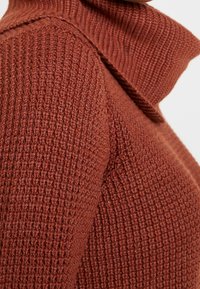 Gros plan sur la manche d'un pull en tricot de couleur rouille et son col à côtes hautes montrant un motif de points texturé.