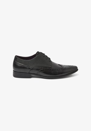 Chaussure habillée pour homme en cuir noir avec perforations en forme d'aile, fermeture à lacets et panneaux latéraux en tissu texturé.