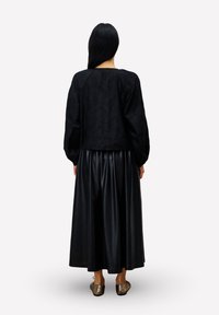 Frau, die mit dem Rücken zur Kamera steht, trägt ein schwarzes Langarmoberteil, einen schwarzen, gefalteten Midirock und metallische Ballerinas vor einem hellen Hintergrund.