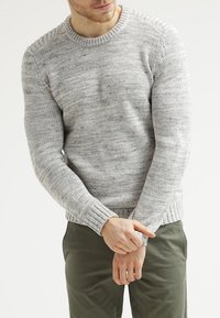 Pull en maille gris clair avec un motif texturé, poignets côtelés et col rond. Porté avec un pantalon vert olive, présentant une coupe décontractée.