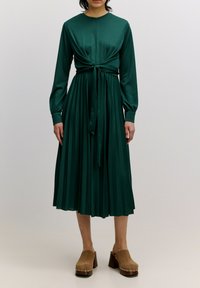 Grünes plissiertes Kleid mit langen Ärmeln, Knoten-Detail an der Taille und rundem Ausschnitt. Kombiniert mit braunen Holzschuhen mit metallischen Akzenten.