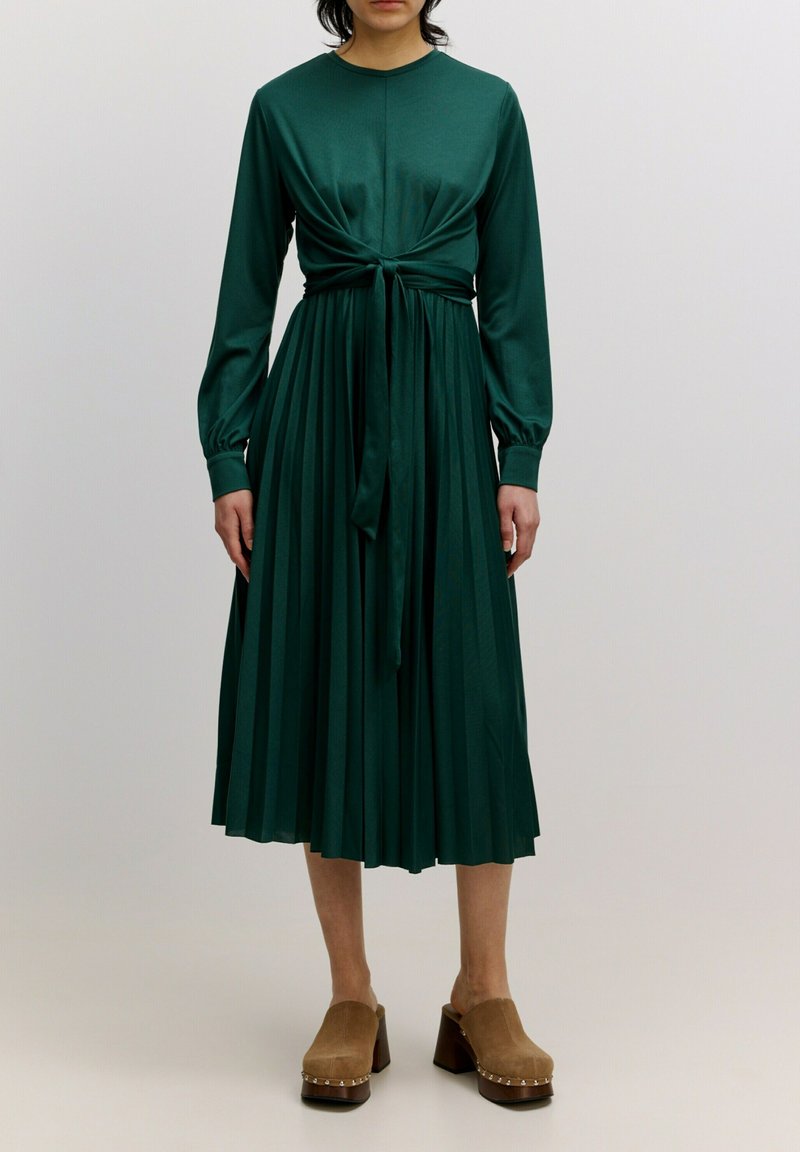 Grünes plissiertes Kleid mit langen Ärmeln, Knoten-Detail an der Taille und rundem Ausschnitt. Kombiniert mit braunen Holzschuhen mit metallischen Akzenten.