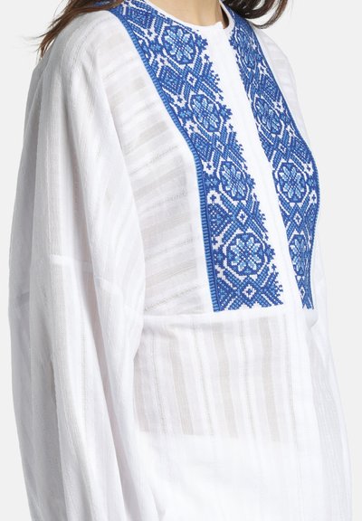 Blouse blanche texturée à manches longues avec une broderie florale bleue verticale sur le panneau de poitrine.