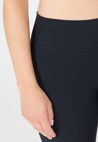 Zwarte high-waisted leggings met een gladde textuur, voorzien van een brede tailleband en platte naden voor comfort. Close-up toont een hand die op de stof rust.