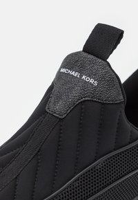 Gros plan d'une basket noire à enfiler avec languette de talon en cuir texturé et logo Michael Kors blanc sur la boucle arrière.