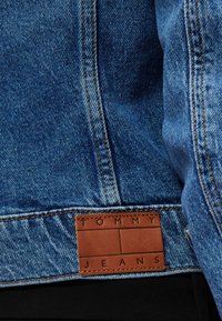 Modrá rifľová bunda s hnedou koženou nášivkou s vyrazeným nápisom "TOMMY JEANS". Viditeľné stehy a mäkká textúra. Klasický dizajn.