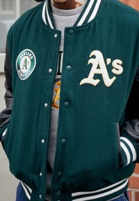 Veste universitaire vert foncé avec manches en cuir noir. Présente un logo des Oakland Athletics et des lettres dorées "A's" sur le devant. Poignets rayés.