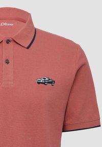 Polo in colore ruggine, realizzato in tessuto testurizzato, con colletto blu navy e una macchina nera ricamata sul petto.