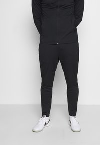 Zwart sportief trainingspak met een ritssluitingjack en taps toelopende broek. Het heeft een gladde textuur en het Nike-logo op de broekspijp, gecombineerd met witte sportschoenen.