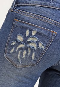 Taisin farkut keskimääräisellä sinisellä pesulla, joissa on kulunut takatasku, jossa on repsottavat reunat ja valkoisia sekä vaaleansinisia brodeerauksia.