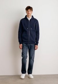 Felpa con zip della marina con cordini, logo anteriore e tasche laterali, abbinata a jeans aderenti in denim scuro e sneakers bianche.
