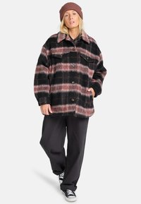 Zwart en roze geruite oversized jas van zacht materiaal, met een kraag, knopen aan de voorkant en twee borstzakken. Draagt bij zwarte broek.