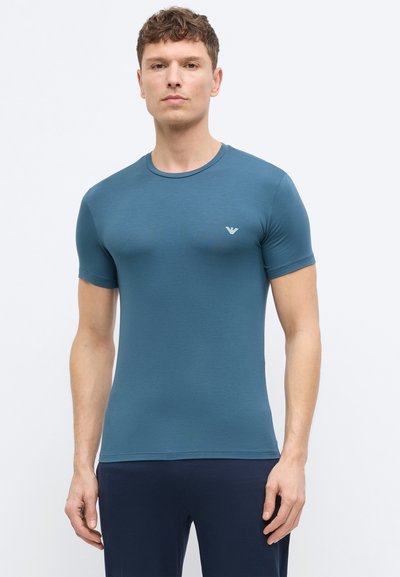 Άνδρας που στέκεται με το σώμα προς τα εμπρός, φορώντας εφαρμοστή πετρόλ κοντομάνικη μπλούζα με μικρό λευκό λογότυπο στο στήθος και σκοτεινό παντελόνι.