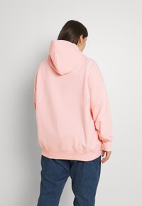 Sudadera rosa con capucha, corte relajado, puños y dobladillo de canalé, combinada con pantalones oscuros azules. Textura suave, diseño minimalista.