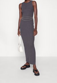 Abito maxi grigio aderente con design senza maniche, accentuato da una cintura in catena argentata. Combinato con sandali platform neri e una borsa bianca.