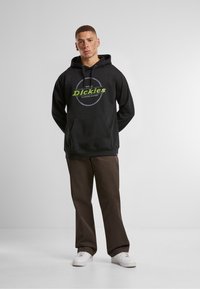 Svart hettegenser med en grønn logo som sier "Dickies" og en sirkulær aksent, sammen med brune bukser og hvite joggesko, på en enkel bakgrunn.
