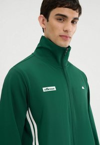 Chaqueta verde con cremallera fabricada en un tejido suave con franjas laterales blancas. Presenta un cuello alto y un pequeño logotipo de la marca en el pecho.