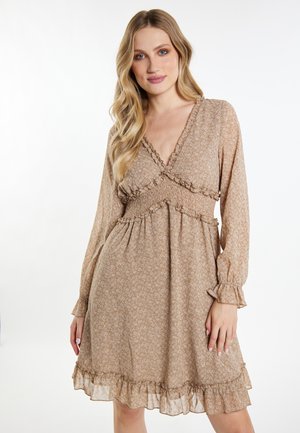 Blonde Frau trägt ein beiges, langärmliges Kleid mit Blumenmuster und Rüschen und steht vor einem weißen Hintergrund.