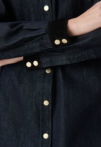 Ciemnoniebieska dżinsowa kurtka z czarnymi aksamitnymi mankietami i złotymi guzikami, noszona z założonymi na klatkę piersiową rękami.