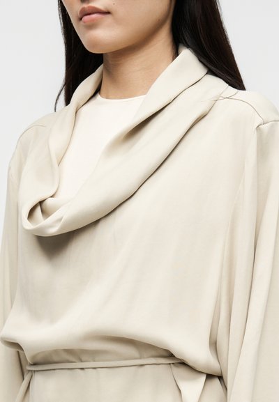 Μπεζ τοπ με λαιμόκοψη φουλάρου από απαλό ύφασμα. Διαθέτει σχεδιασμό με ζάρες και μακριά μανίκια, στενεμένα στη μέση με ζώνη που ταιριάζει.