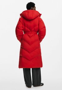 Abrigo largo de color rojo acolchado con capucha, que cuenta con una cintura ceñida por un cinturón y un patrón de acolchado en forma de chevrón. El material parece suave y aislante.