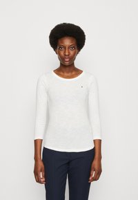 Maglietta bianca a maniche lunghe con scollatura rotonda, caratterizzata da un piccolo logo rosso sul petto, abbinata a pantaloni navy texture.