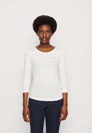 Maglietta bianca a maniche lunghe con scollatura rotonda, caratterizzata da un piccolo logo rosso sul petto, abbinata a pantaloni navy texture.