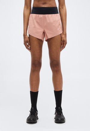 Lichtroze hardloopshorts met een zwarte tailleband, voorzien van een gesplitst ontwerp voor bewegingsvrijheid. Gecombineerd met zwarte sokken en sportschoenen.