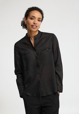 Kobieta o kręconych włosach, nosząca złote kolczyki koła i czarną koszulę na guziki z kieszeniami na piersi, pozująca z rękami w kieszeniach.