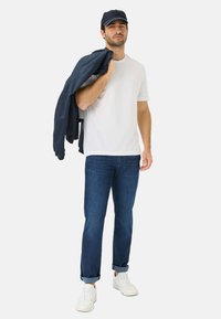 T-shirt bianca, giacca bomber blu scuro appoggiata sulla spalla, jeans blu con i risvolti arrotolati e sneakers bianche, in piedi su uno sfondo neutro.