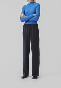 Top lungo a maniche lunghe blu a coste abbinato a pantaloni neri larghi con vita elastica. Il modello si trova in piedi con le braccia incrociate su uno sfondo semplice.