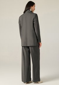 Completo grigio su misura con giacca lunga e pantaloni a gamba larga. Realizzato in tessuto strutturato, presenta dettagli con bottoni sulle maniche della giacca.
