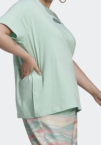 Mentolové tričko s krátkým rukávem vyrobené z měkkého materiálu s kulatým lemem. Výrazné postranní knoflíky a malá černá grafika na přední straně.