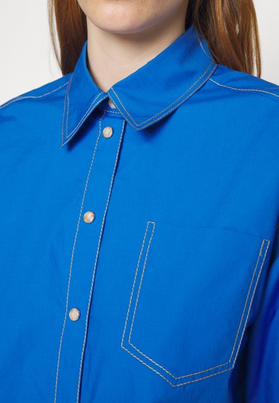 Μπλε πουκάμισο με κουμπιά και αιχμηρό γιακά, με κίτρινη ραφή σε αντίθεση και μία μόνο τσέπη στο στήθος. Λείο υφασμάτινο υλικό.