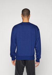 Modra kapuca z dolgimi rokavi, kroja z okroglo vratno otvoritvijo in rebrastimi manšetami. Mehak material in sproščen kroj, prikazan z zadnje strani.